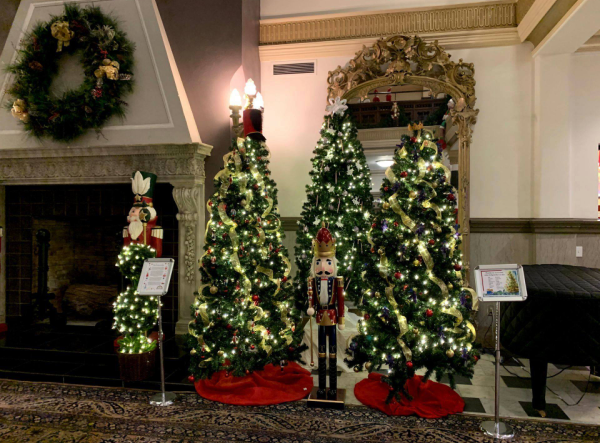 Photograph of three Christmas trees in a group together in the Piqua Public Library lobby. All three trees are decorated with lights and decorations. A large wooden nutcracker stands in front of the middle tree. 