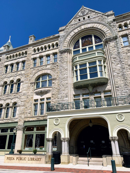 A color photograph of the front of the Fort Piqua Plaza. The image includes the Piqua Public Library sign at the entrance, all the way up to the top of the building. The plaza is a grey stone building with intricate carvings around the windows. The photo depicts a sunny day with a deep blue sky.