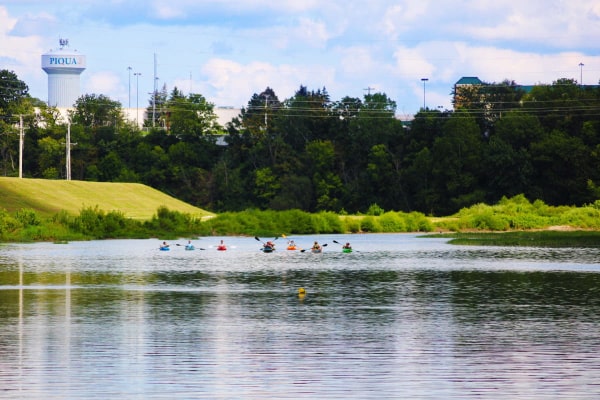 A modern, full-color postcard of the Great Miami River. It's a sunny summer day with blue skies. There are approximately seven kayakers on the water. The Piqua water tower is visible in the background on the left side. The levy is bright green, and the trees along the river are dark green.