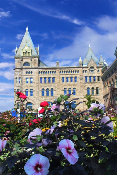 A modern, full-color postcard of the flower gardens off of Main Street in downtown Piqua. Bright green, pink, and red colors are in the foreground. The Fort Piqua Plaza and a deep blue sky make up the background.